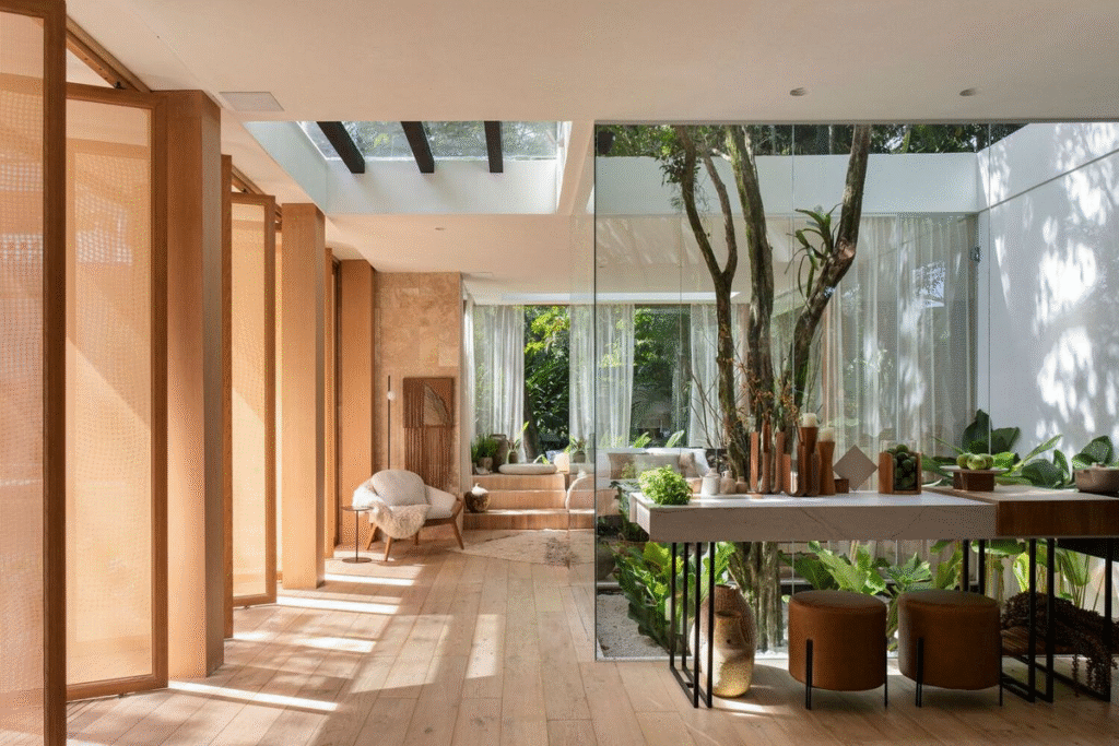 Bright, modern interior featuring warm wooden panels, light oak flooring, and floor-to-ceiling glass walls surrounding an indoor courtyard with mature trees and lush greenery. Natural sunlight streams through a skylight, illuminating a minimalist seating area and a sleek dining table, creating a seamless connection between indoor and outdoor spaces.